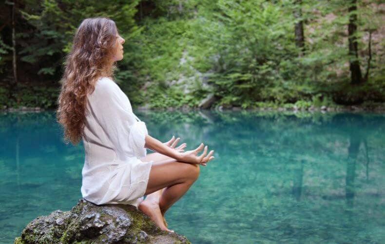 Mulher de perfil meditando em posição de lótus sobre uma rocha, com cabelos longos e vestida de branco, em frente a um lago sereno cercado por árvores verdes.