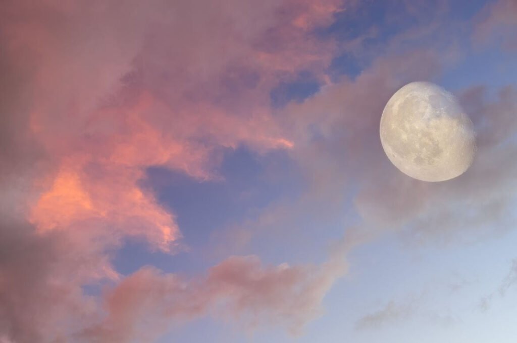 Céu ao entardecer com nuvens tingidas de rosa e laranja e a lua cheia parcialmente visível.