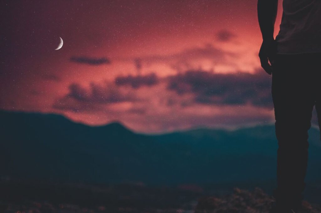 Homem contemplando um céu noturno estrelado sobre montanhas com lua crescente.