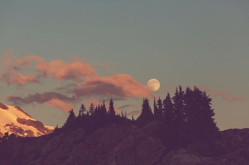 Lua cheia aparecendo sobre pinheiros e montanhas ao entardecer com nuvens cor de rosa e céu em tons pastel