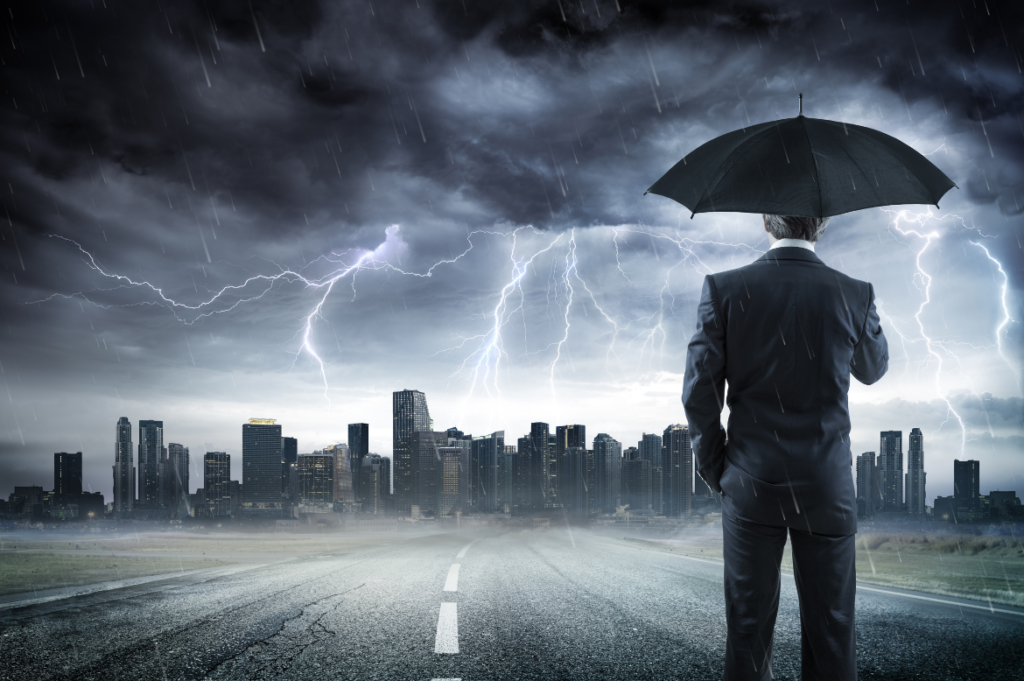 Imagem de um homem de terno segurando um guarda-chuva, olhando para uma cidade ao fundo sob um céu tempestuoso. Relâmpagos cortam o céu escuro, e a chuva cai intensamente, criando uma atmosfera de tensão e desafio. A estrada molhada e a paisagem urbana envolta em nuvens escuras sugerem uma jornada difícil à frente, simbolizando resiliência em meio a adversidades. Ideal para conteúdos sobre superação, gestão de crises, perseverança no ambiente corporativo e preparação para desafios econômicos ou profissionais.